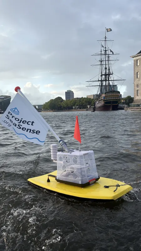 The SeaSense prototype on its yellow board with a Project SeaSense flag, on the water in Amsterdam with a tall ship behind it